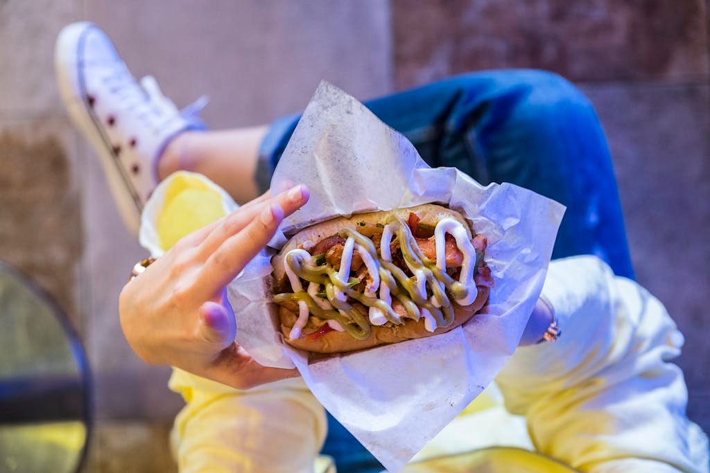 A person holding a delicious hot dog with toppings, captured from above outdoors in a vibrant setting.