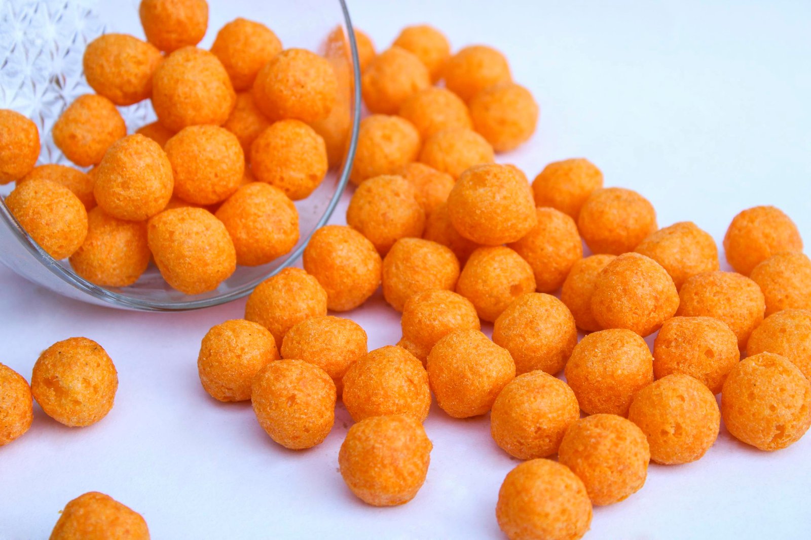 A close-up of orange cheese balls spilling from a glass bowl on a white background.