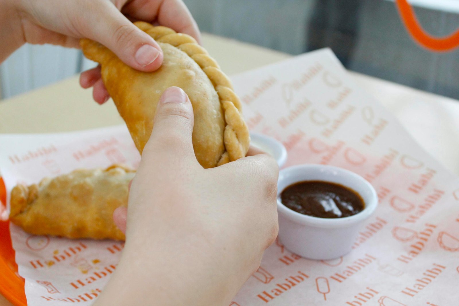 A person holding a freshly baked empanada with dipping sauce on a tray.