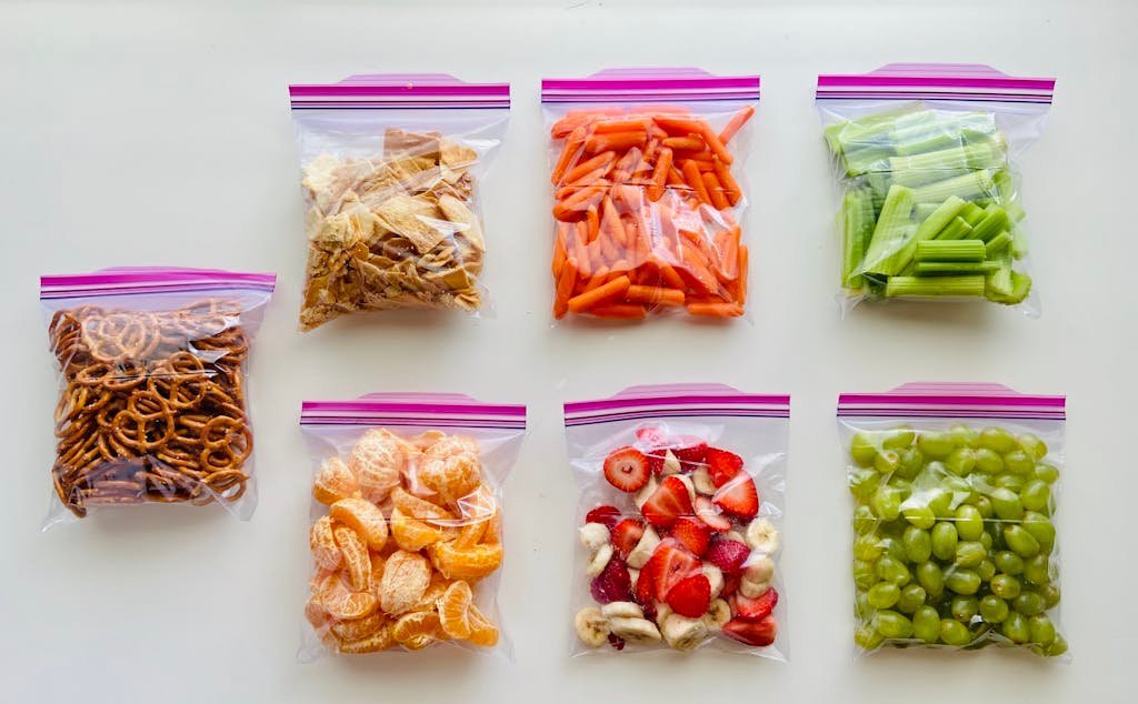 An assortment of fruits, vegetables, and snacks neatly packed in zip lock bags on a white background.