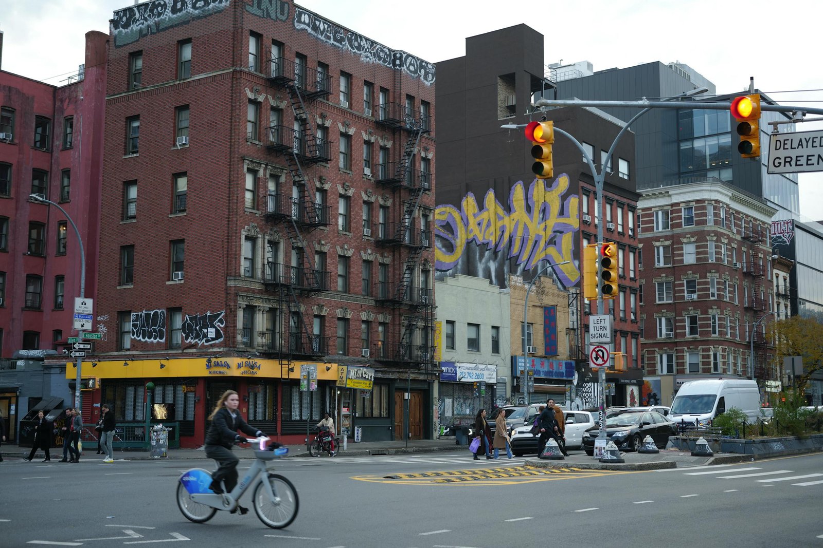 Busy urban intersection with cyclist, pedestrians, and graffiti-laden buildings in cityscape.